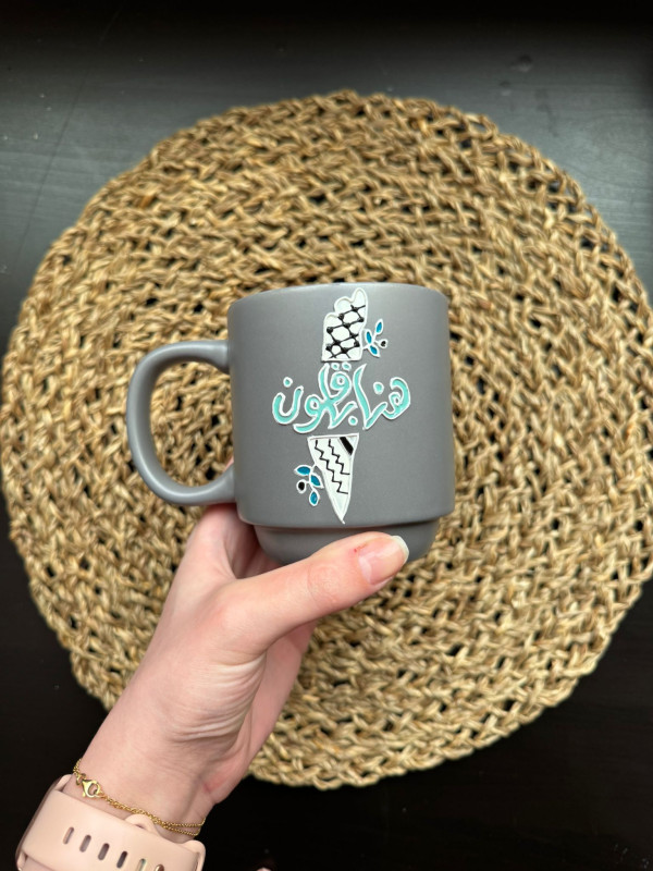 A gray cup with a written phrase on it / we remain (Palestine) made by hand